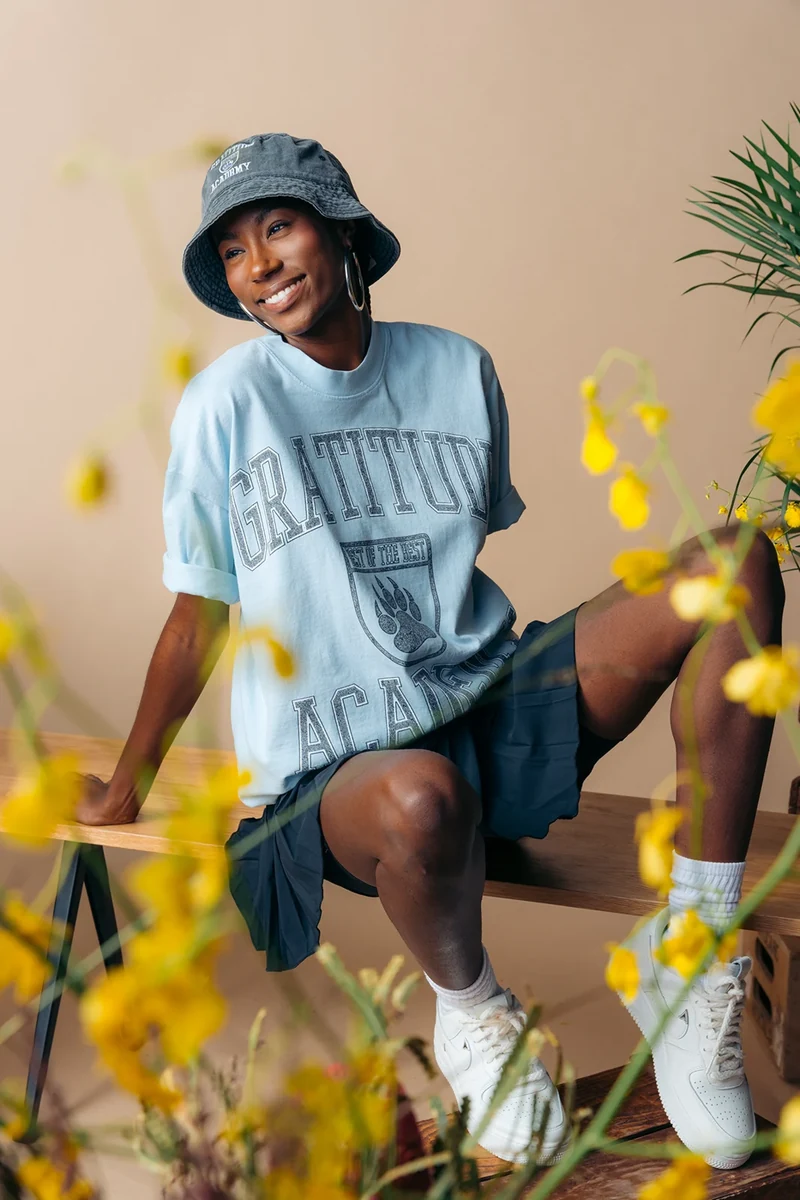 Seated model in a blue Gratitude Academy tee, framed by yellow flowers.
